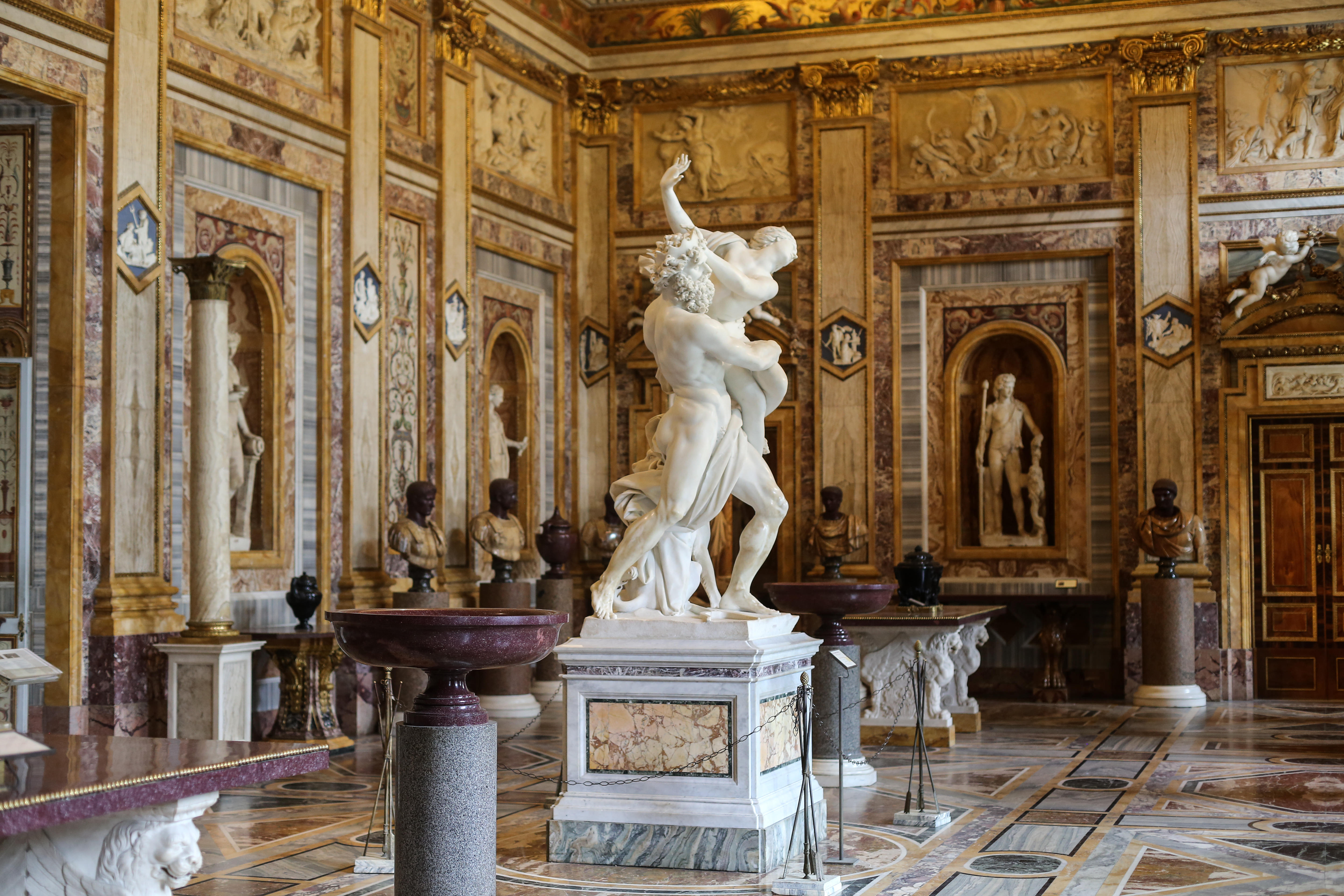 A large marble statue of a male figure grabbing a female stands in the center of a gallery