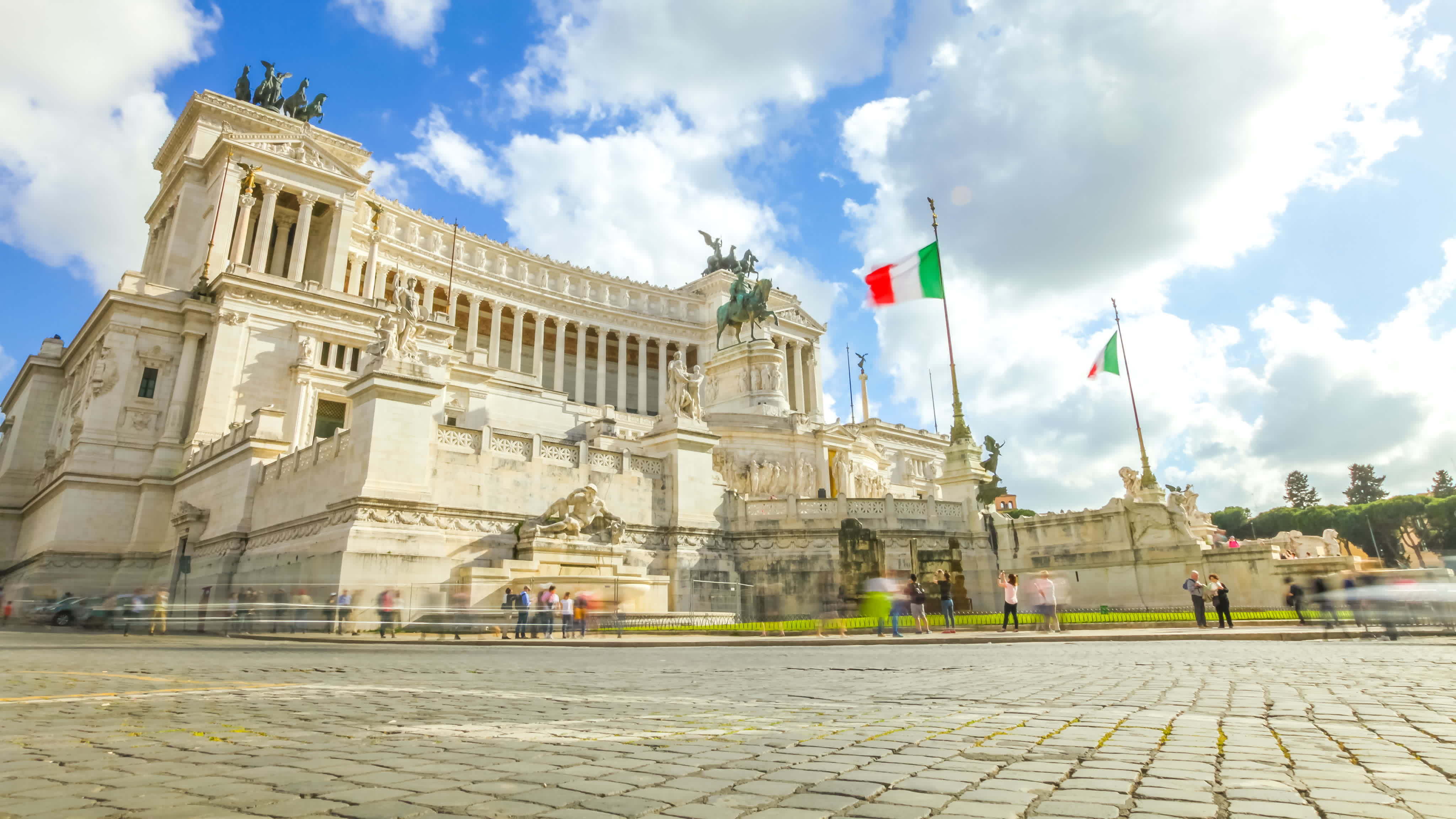 A massive white building surrounded by many statues with Italian flags flying outside