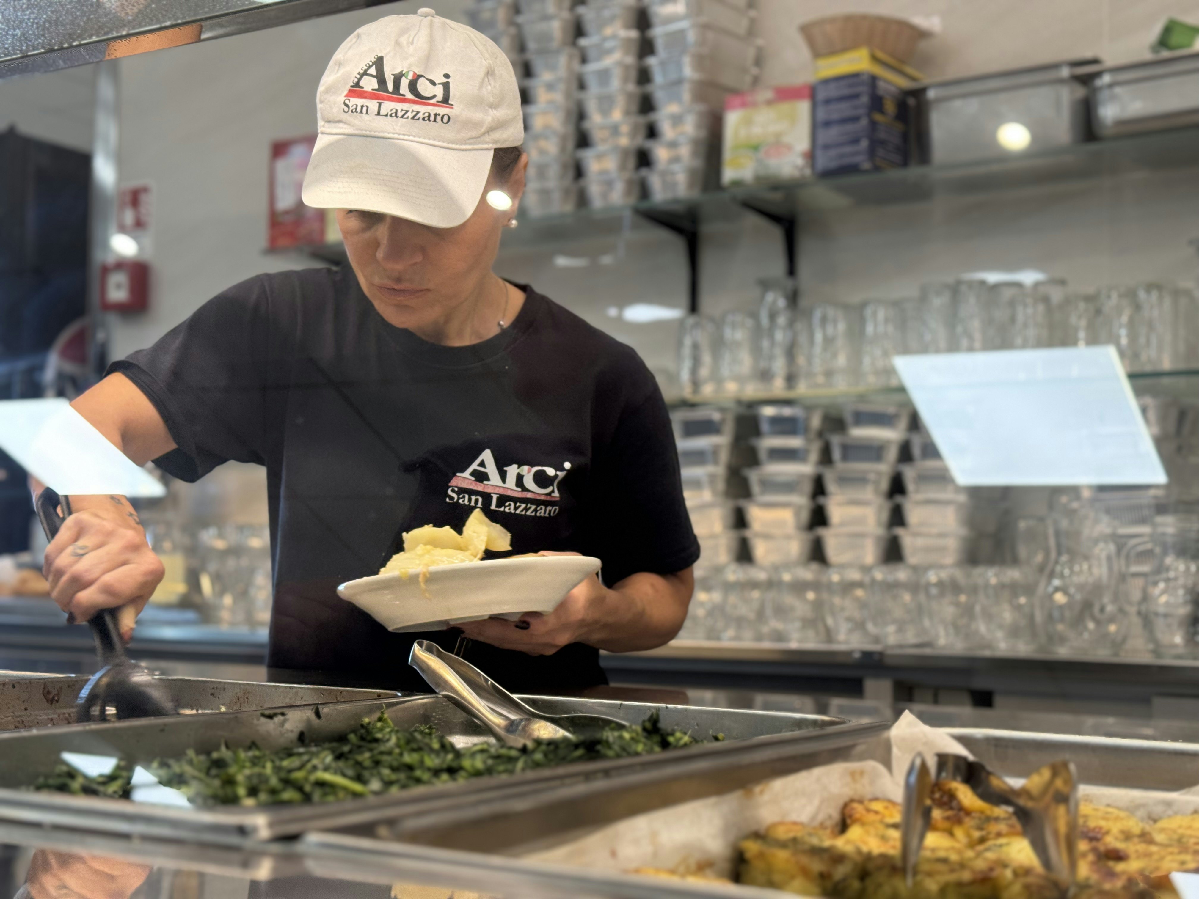 A woman in a dark blue T-shirt and a white cap serves food onto a white plate from a buffet warmer line
