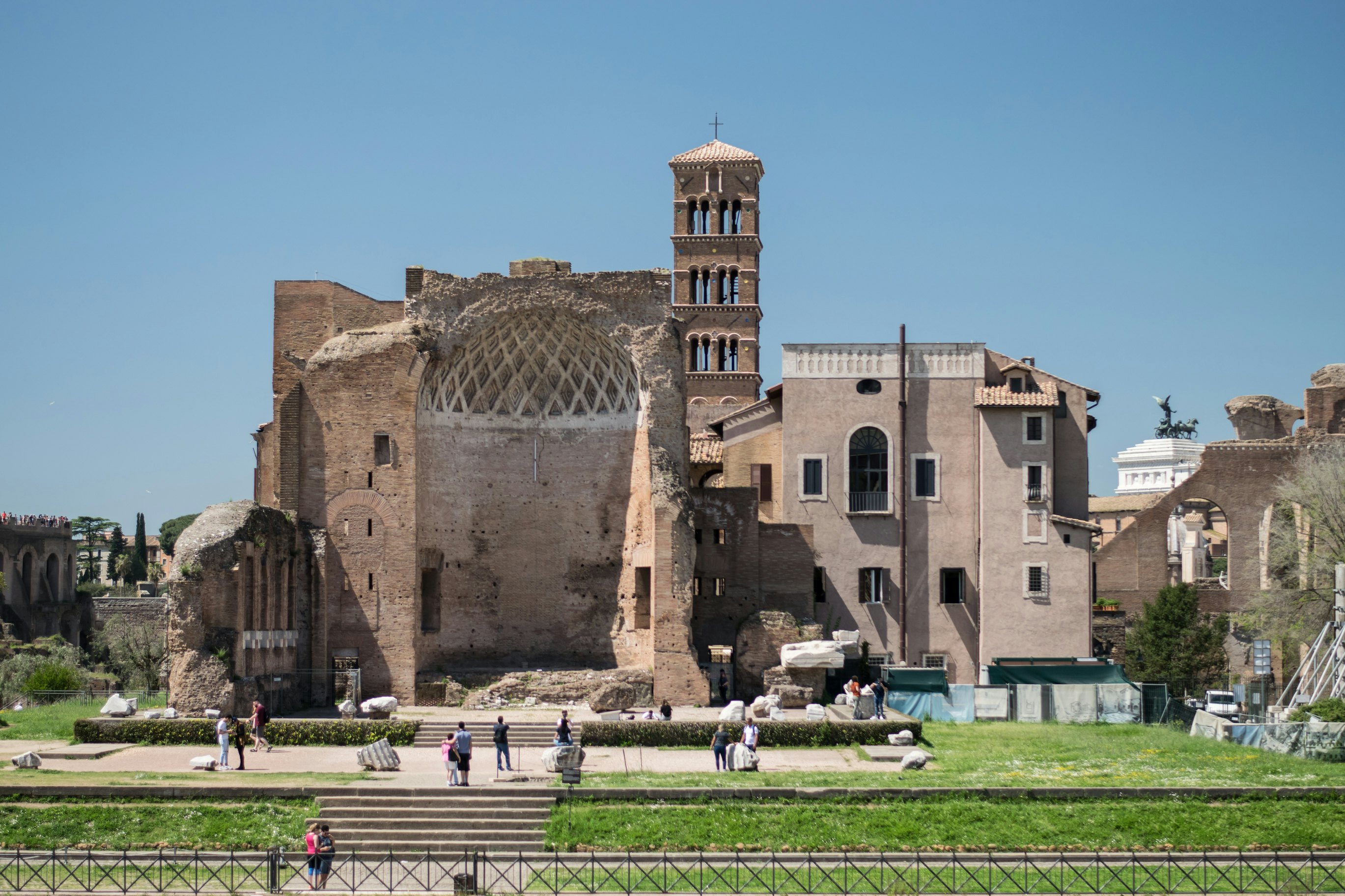 Tourists gather in a park to view the Ancient Roman ruins of a grand villa, one part of the wall is missing so that the interior and dome-shaped roof is visible.