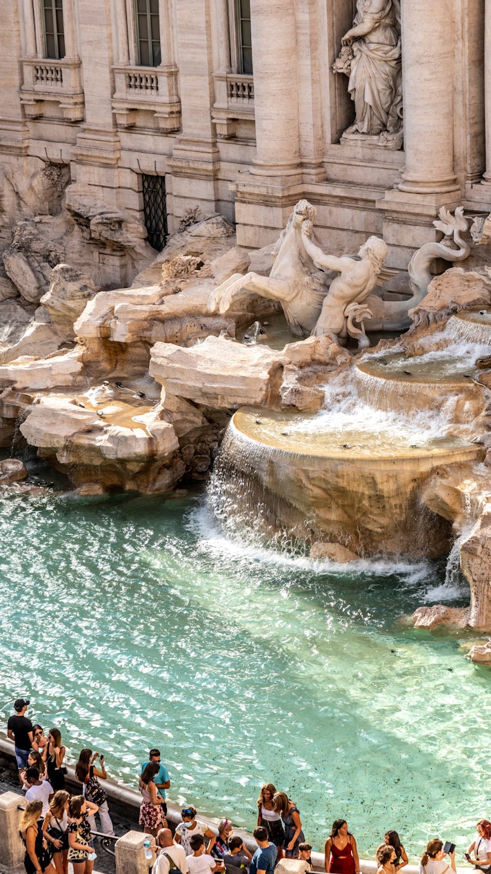 People gather in front of the clear waters of the Trevi fountain, right in front of the giant marble statues.