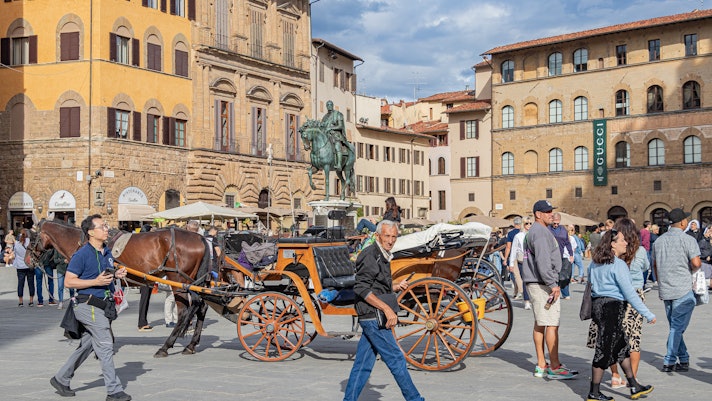 A crowd of people walk through a square framed by museums. Horse-drawn carriages are waiting for passengers.