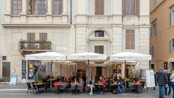 People dine under parasols at an outdoor terrace of a white-washed stone building.